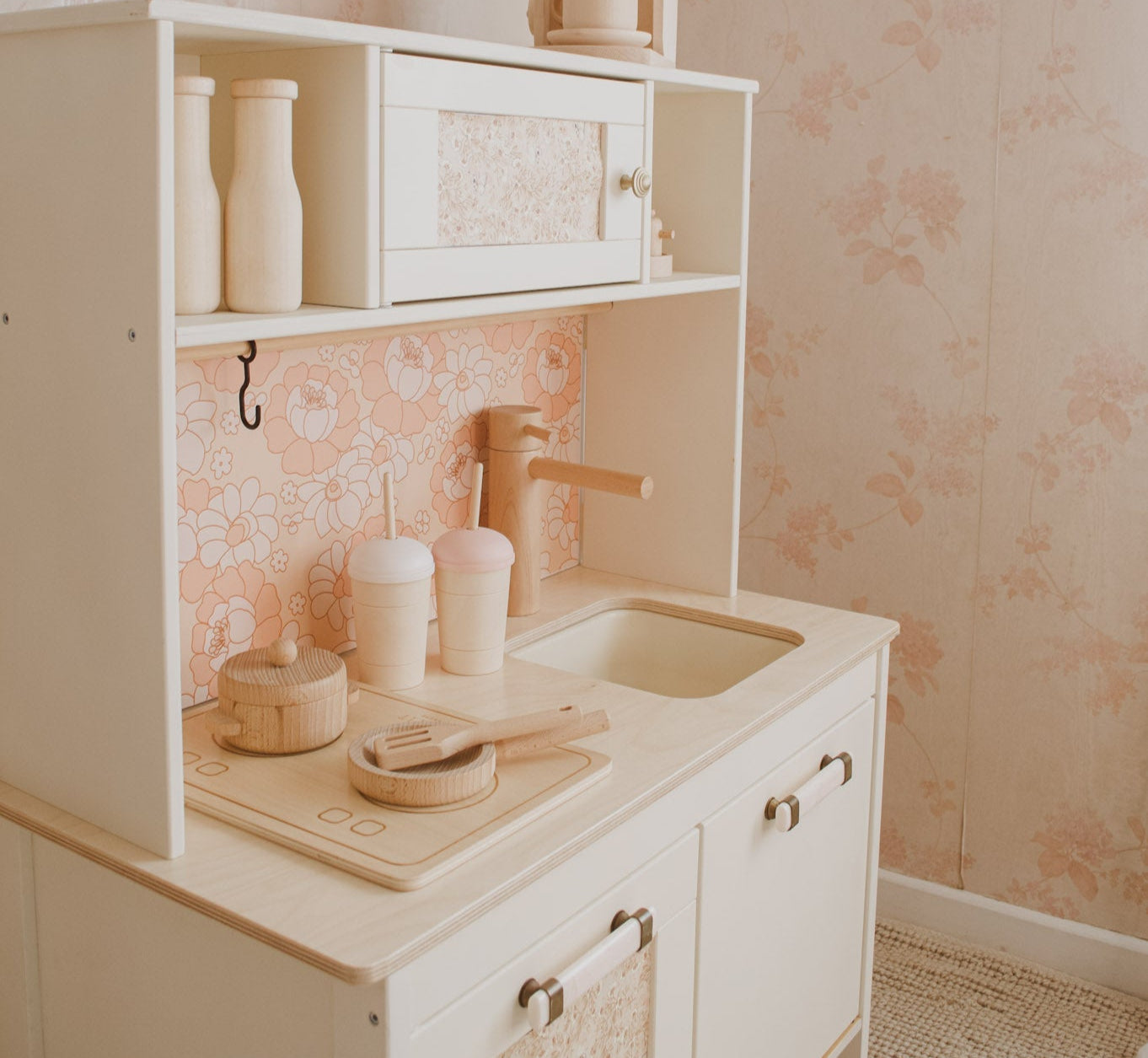 Children's play kitchen set with sink and utensils against a floral wallpapered wall.